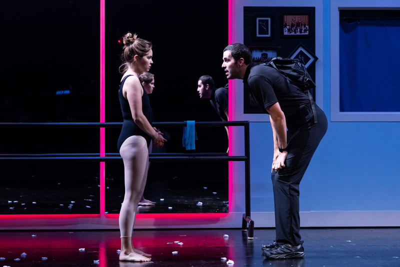 a young woman stands on a stage in a leotard and tights stands facing a man who bent down looking at her and speaking to her.
